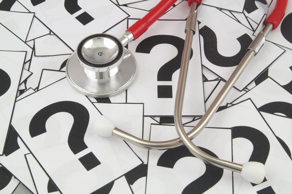 Image of a pile of papers with just a single question mark on each. Laying on top of the papers is a silver and red stethoscope.